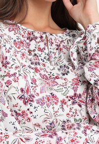 Blumenbluse mit weißem Hintergrund, die rot, blau und grün blühende Blumen zeigt, einem runden Ausschnitt und einem kleinen Schlüsselelement.