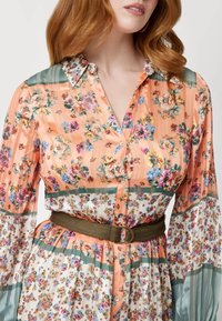 Gėlių raštas mygtukais užsegamas suknelė su oranžinėmis ir žaliosiomis horizontaliosiomis juostelėmis, turinti ilgus rankovėmis, apibrėžtą juosmenį su chaki diržu ir šilkine tekstūra.