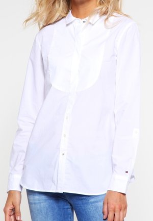 Kobieta ubrana w białą koszulę na guziki z długim rękawem i zaokrąglonym przodem oraz jasnoniebieskie dżinsy.