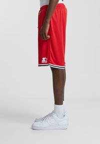 Pantaloni scurți atletici roșii cu talie cu șnur, margine cu dungi negre și albe și un logo în colțul din stânga jos. Asortați cu pantofi sport albi.