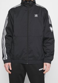 Svart sportjacka med vit tre-streksdesign på ärmarna, hög krage, framficka med dragkedja och broderad logotyp på bröstet och ärmen.