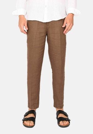 Uomo che indossa pantaloni marroni a taglio slim, sandali neri con fibbia e una camicia bianca a maniche lunghe, in piedi davanti a uno sfondo semplice.