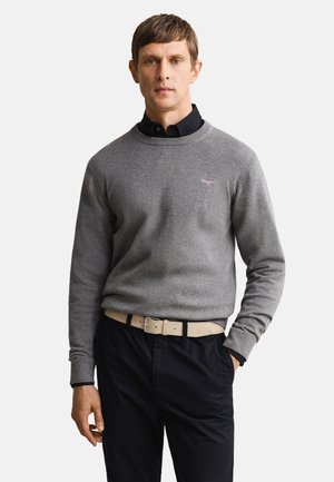 Mand iført grå sweater over sort skjorte med krave, beige bælte og sorte bukser, der står med den ene hånd i lommen mod hvid baggrund.