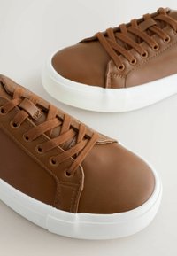 Ruske nahkakengät valkoisilla kumipohjilla, joissa on litteät ruskeat nauhat ja metalliset silmäkappaleet. Sileä pinta ja pyöristetty kärkitoteutus.