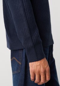 Navy ribbed sweater with a textured knit, featuring a close-fitting cuff. Worn with dark denim jeans, showing a detail of the hand.