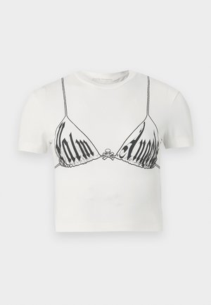 Γυναίκα που φοράει λευκό cropped t-shirt με γραφικό σουτιέν, πράσινη και ναυτικό πλισέ φούστα, λευκές κάλτσες και μπεζ και μαύρα αθλητικά παπούτσια, όρθια.