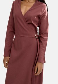 Sukienka kopertowa w burgundowym kolorze z długimi rękawami, dekoltem w serek oraz bocznym paskiem z detalem w postaci srebrnego pierścienia. Gładka faktura, dopasowany krój.