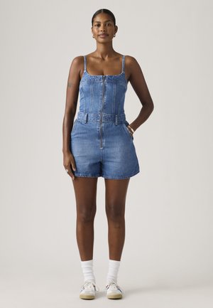 Žena stojaca, oblečená v bezrukávovom rifľovom overal s predným zipsom, bielymi ponožkami a bielymi teniskami na jednofarebnom pozadí.