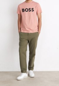 Růžové bavlněné tričko s nápisem "BOSS" tištěným tučným černým písmem, v kombinaci s olivově zelenými bavlněnými kalhotami a bílými teniskami.