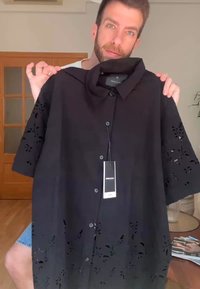 Μαύρο κοντομάνικο πουκάμισο με ανθρωπομορφικούς κοπής στο τελείωμα, κουμπιά μπροστά και ετικέτα προσαρτημένη, κατασκευασμένο από ελαφρύ ύφασμα.