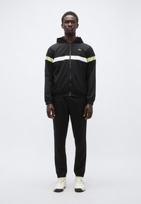 Musta vetoketjullinen takki, jossa vihreät ja valkoiset raidat, yhdistetty mustiin housuihin ja valkoisiin tennareihin. Takissa on huppu ja joustavat hihansuut.