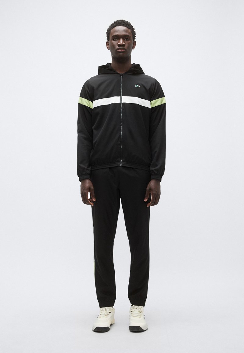 Musta vetoketjullinen takki, jossa vihreät ja valkoiset raidat, yhdistetty mustiin housuihin ja valkoisiin tennareihin. Takissa on huppu ja joustavat hihansuut.