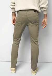 Man in beige chinos, crème sweater en beige sneakers, met zijn rug naar de camera gekeerd tegen een effen witte achtergrond.