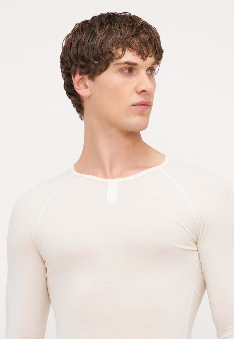 Camicia a maniche lunghe color beige chiaro con scollo rotondo, realizzata in un tessuto liscio e elasticizzato. Presenta un piccolo etichetta nell'area del colletto.