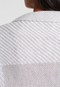 Lehký, texturovaný pletený top v jemné béžové a krémové barvě se pruhovaným vzorem a klasickým límečkem, který zobrazuje otevřené tkaní a detaily.