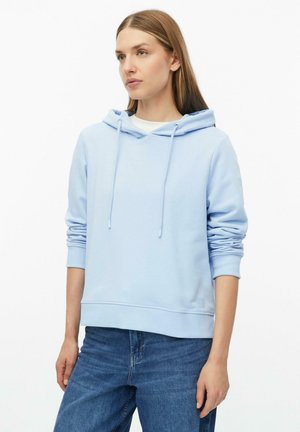 Frau trägt hellblauen Kapuzenpullover mit Kordelzug und blaue Jeans und steht vor weißem Hintergrund.