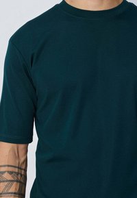 T-shirt à manches courtes en teal foncé, fabriqué en coton doux. Encolure ronde et design simpliste sans motifs ni accents visibles.