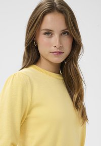 Pull jaune avec un col rond, bord côtelé et manches légèrement bouffantes. Texture douce et coupe ajustée. Légère brillance.