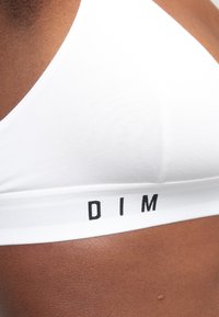 Bralette blanca de algodón con forma triangular y textura suave, con un logotipo negro "DIM" en la banda elástica.