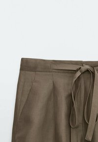 Brown linen trousers featuring a high waist, pleats, and a tied belt. The fabric appears smooth with a subtle texture.