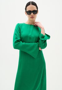 Zöld selyemruha hosszú harangujjakkal, egyszerű kerek nyakkivágással és sima textúrával. A modellen fekete napszemüveg van.
