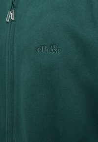 Tmavě zelená zipová bunda vyrobená z jemné tkaniny, s vystouplým logem "ellesse" na hrudi. Detaily táhla zipu dodávají jemné akcenty.