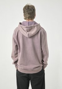 Pullover-Hoodie aus weichem, gedämpftem Lavendelstoff mit einer Kordelzugkapuze, gerippten Bündchen und Saum. Getragen mit schwarzen Hosen, die einen lockeren Schnitt haben.