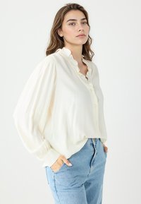 Jasna kremowa bluzka na guziki z falbaniastym kołnierzykiem i długimi rękawami. Miękki, lekki materiał o luźnym kroju, w połączeniu z niebieskimi dżinsami.