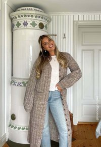 Brede, lange cardigan in lichtbruin met ribbelstructuur, voorzakken en knopen; gecombineerd met lichtblauwe jeans en een witte blouse.