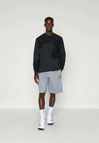 Muž na sobě má černou mikinu, světle šedé šortky, bílé ponožky Nike a bílé tenisky Nike, stojí na jednobarevném pozadí.