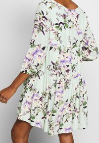 Jasno miętowa sukienka z rękawami do trzech czwarte, w kwiatowy wzór z fioletowymi i żółtymi kwiatami, miękka tkanina, warstwowy krój i zaokrąglony dekolt.
