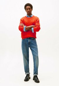 Maglione arancione a trecce sopra una camicia button-up azzurra chiara, abbinato a jeans azzurri chiari con risvolti e scarpe nere.