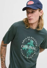 Mørkegrøn bomulds t-shirt med et cirkulært blomstermotiv og teksten "ARIA ELEGANCE ORIGINALS." Modellen bærer en marineblå cap.