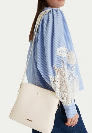 Mujer con blusa de rayas azules y blancas con detalles de encaje, llevando un bolso de hombro color crema con logo dorado y correa ajustable.