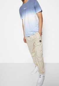 Camiseta de color azul claro con degradado y logo, pantalones cargo beige con bolsillos laterales y zapatillas blancas con detalles en negro. Un atuendo simple y casual.