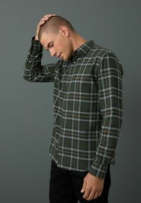 Chemise à carreaux verte à boutons, avec des lignes grises et jaunes, fabriquée dans un tissu à texture douce. La chemise a un col classique et des manches longues.