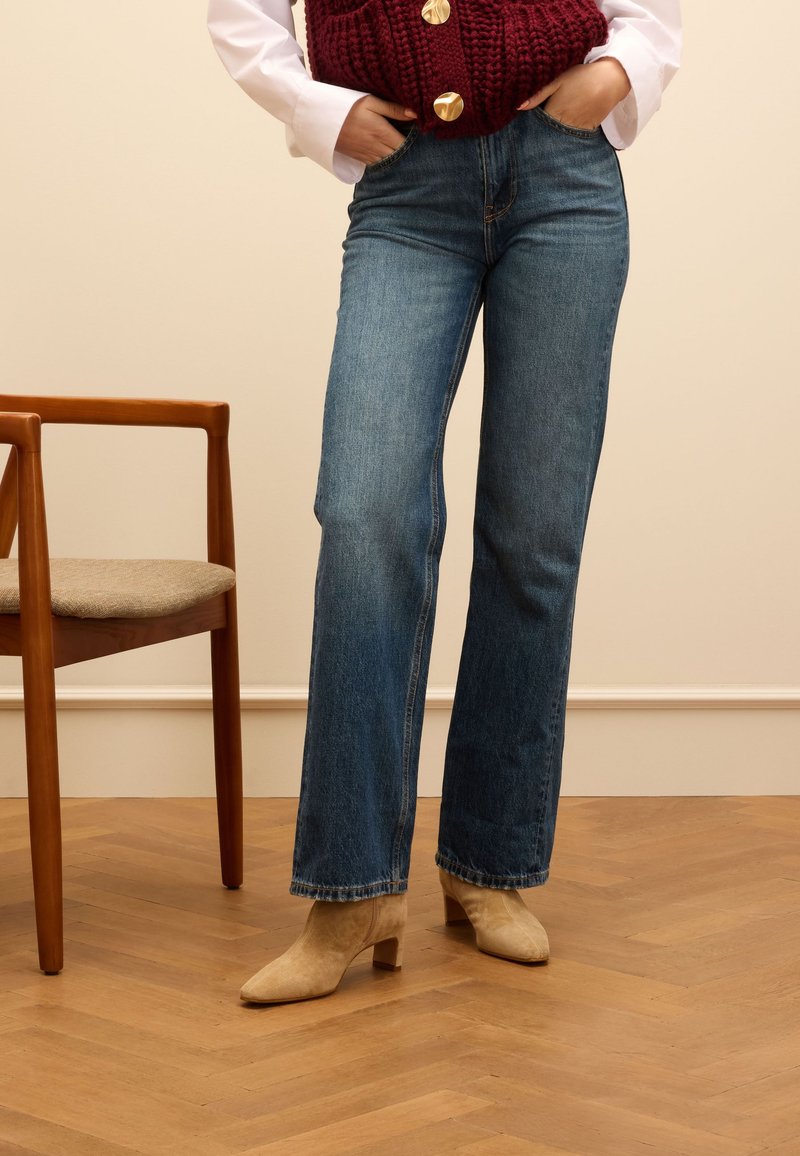 Blaue, ausgestellte Jeans aus Denim mit hoher Taille, klassischem Fünf-Taschen-Design und einem hellen Waschung. Kombiniert mit beigen, high-heeled Stiefeletten.