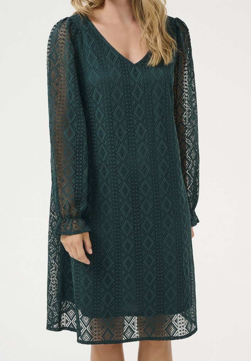 Femme portant une robe vert foncé jusqu’aux genoux avec des manches longues en dentelle et un col en V, mettant en valeur un motif géométrique en dentelle.