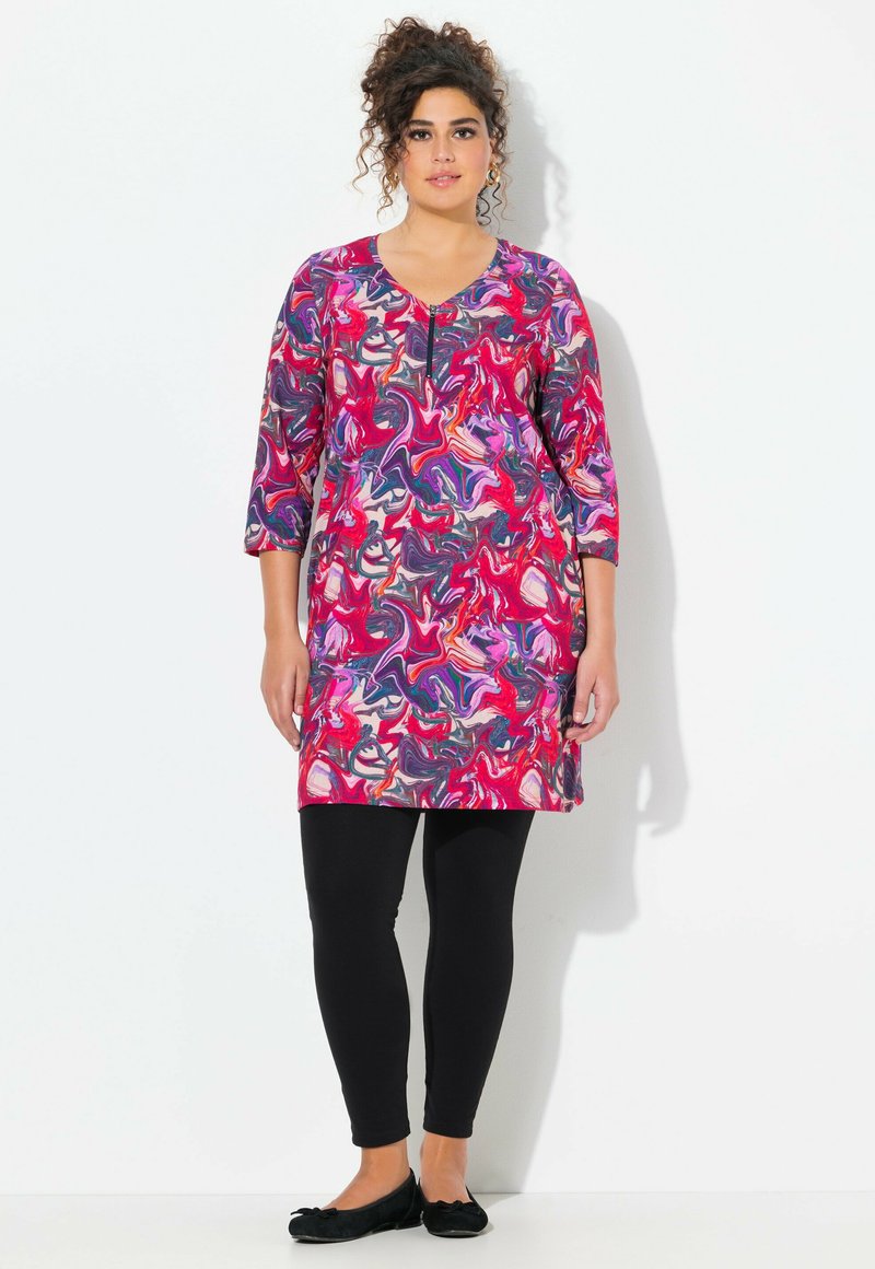Kleurige tuniek met draaiende roze, paarse en witte patronen; driekwart mouwen; gecombineerd met zwarte leggings en platte zwarte schoenen.