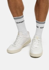 Białe skórzane trampki z perforowaną przednią częścią i jasną podeszwą, w zestawieniu z białymi skarpetkami z czarnymi paskami i napisem z logo.