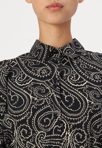 Czarna bluzka w wzór z paisley w kolorze kremowym. Krótki kołnierzyk, guziki z przodu oraz strukturalny materiał.