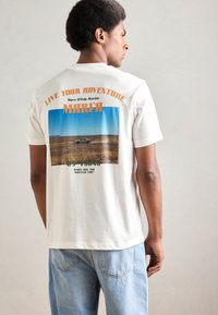 Weißes Baumwoll-T-Shirt mit einem grafischen Druck auf der Rückseite, der Text und ein Landschaftsbild zeigt, kombiniert mit hellblauen Jeans.