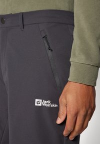 Sorte softshell bukser har en lynlåslomme, logo-patch og flad talje. Det teksturerede stof giver stræk og holdbarhed.