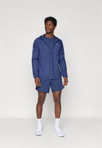 Άνδρας που στέκεται σε ναυτικά μπλε αθλητική ζακέτα, αντίστοιχα σορτς, λευκά κάλτσα και λευκά αθλητικά παπούτσια σε απλή ανοιχτόχρωμη φόντο.