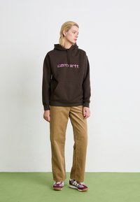 Bruine hoodie met roze logo, voorzak en capuchon met trekkoord. Gecombineerd met tan corduroy broek en bordeauxkleurige sneakers met witte accenten.