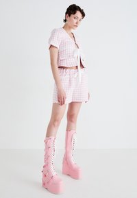 Veste courte en tweed rose avec un nœud à l'avant et un short assorti, associée à des bottes plateformes roses montantes ornées de chaînes décoratives.