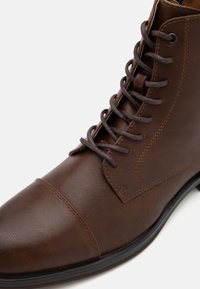 Koffiekleurig leren enkelboot met een ronde neus, vetersluiting aan de voorkant en minimale stikseldetails. Heeft een gladde textuur en een stevige zool.
