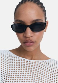 Czarne okulary przeciwsłoneczne w kocim kształcie z eleganckim, minimalistycznym designem. Wykonane z plastiku, posiadają przyciemnione soczewki i gładkie wykończenie. Noszone z topem zrobionym na szydełku.