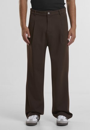 Mann trägt lockere dunkelbraune Hose und weiße Turnschuhe, steht vor einem schlichten hellen Hintergrund.