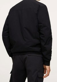Veste bomber noire avec une finition lisse et mate, présentant une coupe décontractée, des poignets côtelés et un design épuré sans marque visible.
